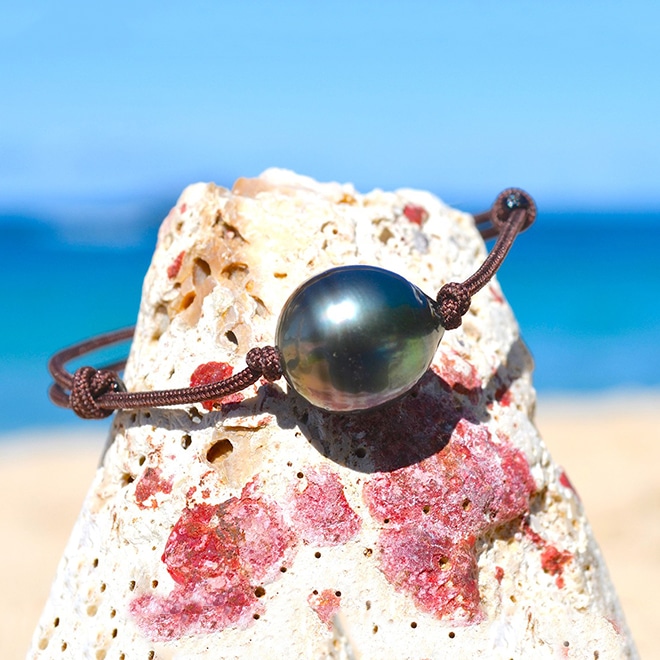 Adjustable brown cord bracelet with a 13 mm circled Tahitian cultured pearl displayed on a rock by the sea