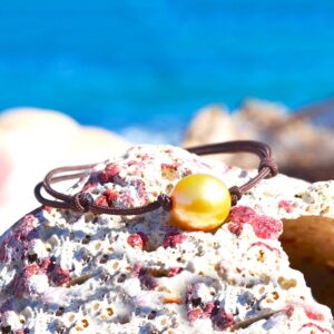 Bracelet ajustable sur cordon marron avec une perle de culture dorée d’Australie de 11,5 mm présenté sur une roche face à la mer