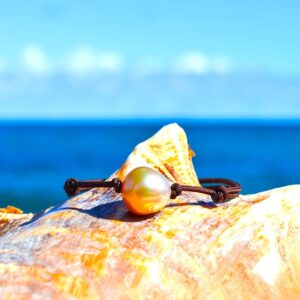 Bracelet ajustable sur cordon marron avec une perle de culture dorée d’Australie de 12 mm présenté sur un coquillage face à la mer