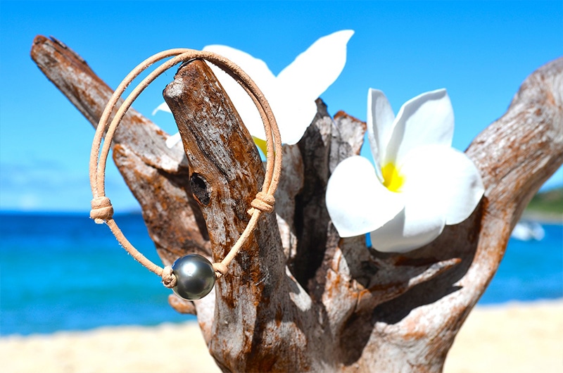 Adjustable leather bracelet with a 12.5 mm round Tahitian pearl displayed on wood with a white flower by the sea