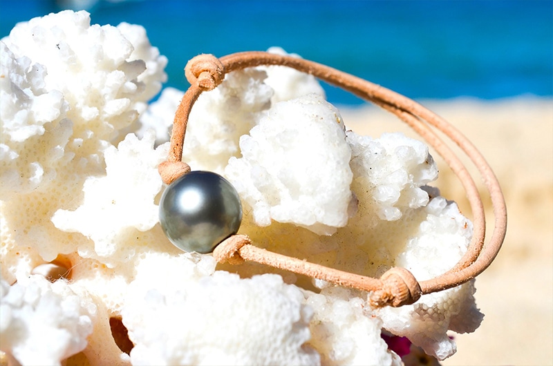 Adjustable leather bracelet with a 12.5 mm round Tahitian pearl displayed on coral by the sea