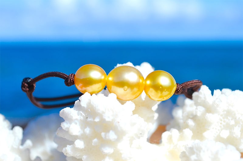 Bracelet ajustable avec trois perles dorées d’Australie de 8,5 mm présenté sur du corail blanc face à la mer