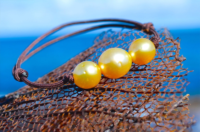 Bracelet ajustable avec trois perles dorées d’Australie de 8,5 mm présenté sur un filet face à la mer