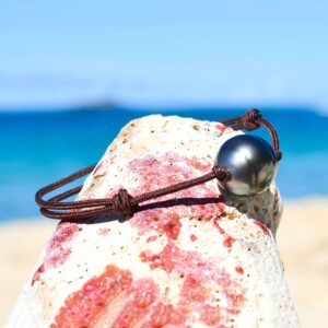 Adjustable brown cord bracelet with a 12.5 mm circled Tahitian cultured pearl displayed on a rock by the sea