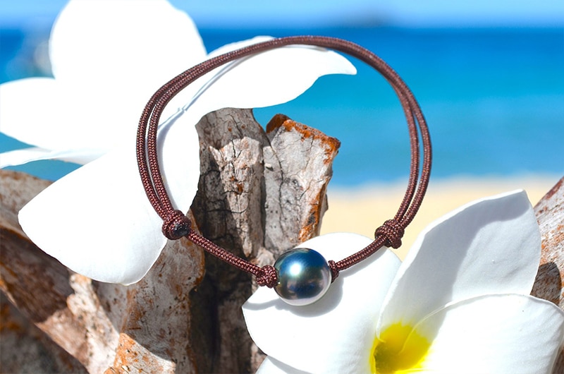 Bracelet ajustable avec une perle semi-ronde de Tahiti de 9 mm présenté sur du bois avec des fleurs blanches face à la mer