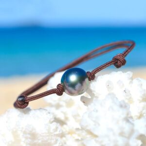 Adjustable brown cord bracelet with a 9 mm semi-round Tahitian cultured pearl displayed on white coral by the sea