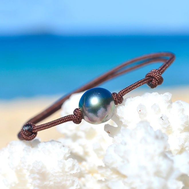 Bracelet ajustable sur cordon marron avec une perle de culture de Tahiti semi-ronde de 9 mm présenté sur du corail blanc face à la mer