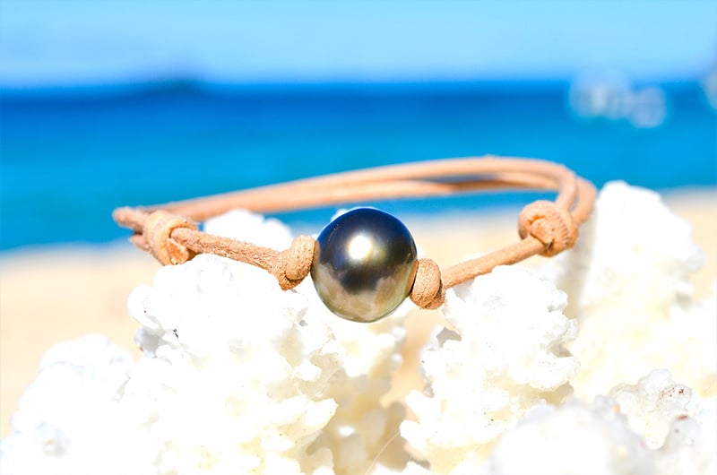 Adjustable leather bracelet with a 12 mm semi-round Tahitian pearl displayed on white coral by the sea