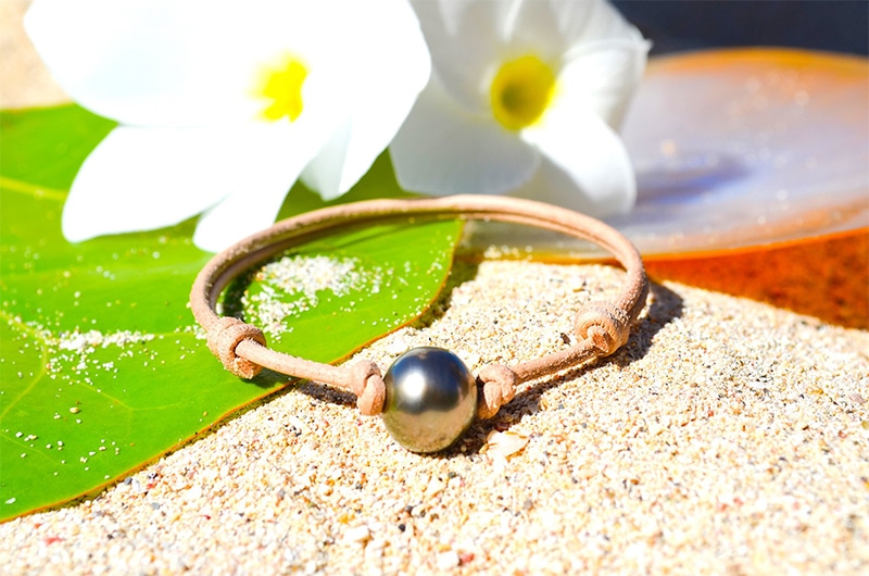 Full view of the adjustable leather bracelet with a 12 mm semi-round Tahitian pearl displayed on a leaf with white flowers