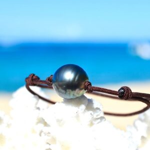 Adjustable brown cord bracelet with a 12 mm circled Tahitian cultured pearl displayed on white coral by the sea