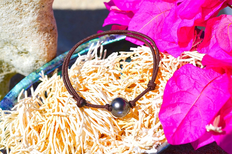Adjustable brown MOKUBA cord bracelet with a 9.5 mm semi-round Tahitian cultured pearl displayed on a shell with pink flowers