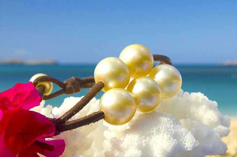 Vue d’ensemble du bracelet en cuir avec cinq perles dorées d’Australie présenté sur du corail blanc face à la mer
