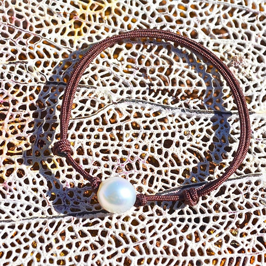 Adjustable brown cord bracelet with a white Australian cultured pearl, laid flat on coral
