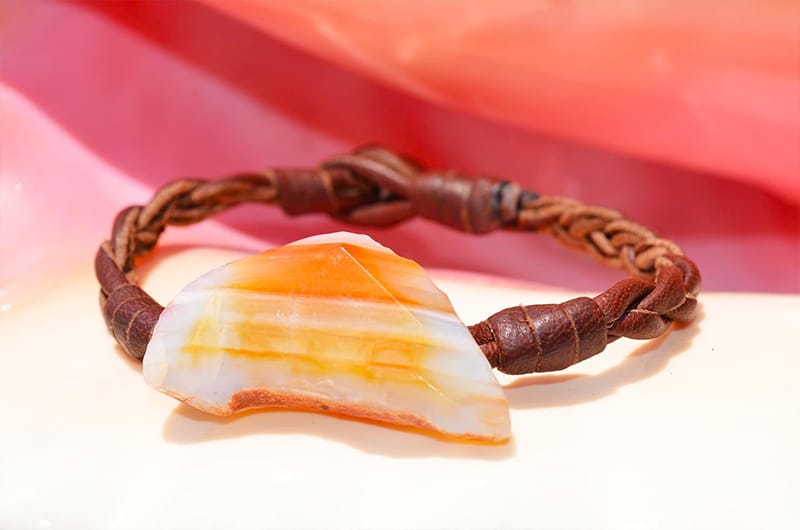 Full view of the braided leather bracelet with an orange agate and a Tahitian pearl displayed on a pink background