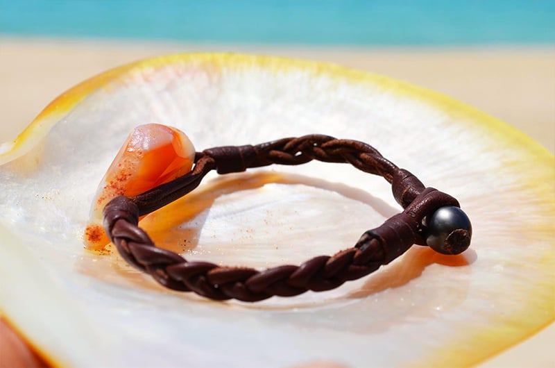 Braided leather bracelet with an orange agate and a Tahitian pearl displayed on mother-of-pearl by the sea