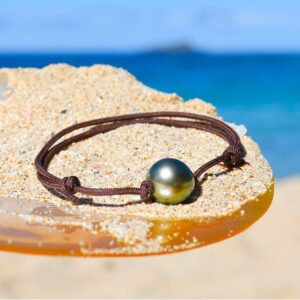 Adjustable brown cord bracelet with a 10.5 mm semi-round Tahitian cultured pearl displayed on sand by the sea