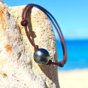 Full view of the adjustable bracelet with a 10 mm semi-round Tahitian pearl displayed on a rock by the sea