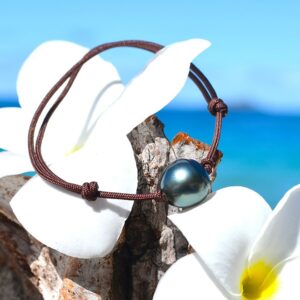 Adjustable brown MOKUBA cord bracelet with an 11.5 mm semi-baroque Tahitian cultured pearl displayed on wood with white flowers by the sea