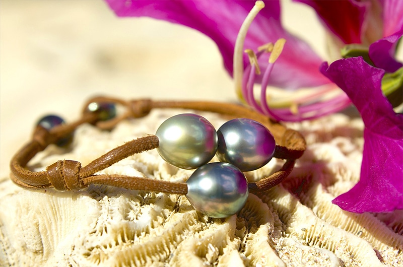 Vue d’ensemble du bracelet en cuir avec trois perles de Tahiti présenté sur un coquillage avec des fleurs roses