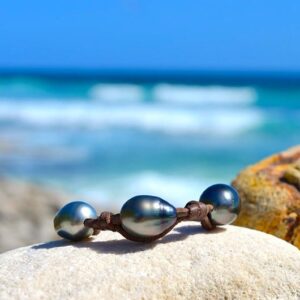 Full view of the leather bracelet with three 11.5 mm Tahitian pearls and knots displayed on a rock by the sea