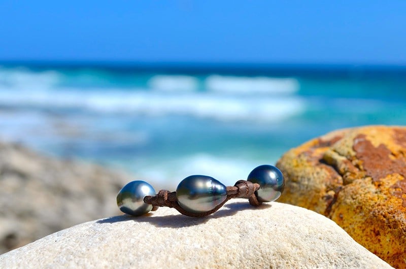 Full view of the leather bracelet with three 11.5 mm Tahitian pearls and knots displayed on a rock by the sea