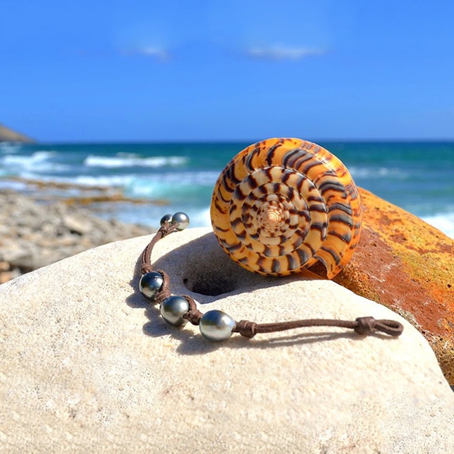 Brown leather bracelet with three central 11.5 mm Tahitian pearls, knots and two closure pearls displayed on a rock with a shell by the sea