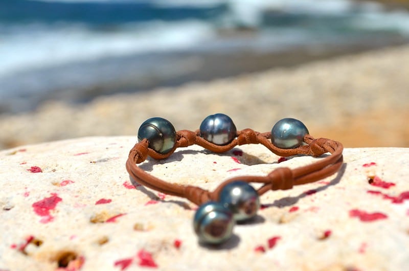 Full view of the leather bracelet with three 11 mm circled Tahitian pearls and knots displayed on a rock by the sea