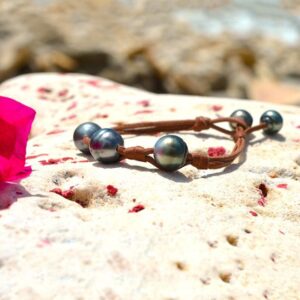 Brown leather bracelet with three central 11 mm circled Tahitian pearls, knots and two closure pearls displayed on a rock with a pink flower by the sea