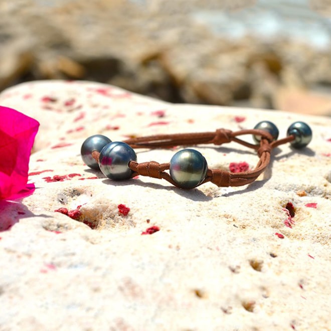 Brown leather bracelet with three central 11 mm circled Tahitian pearls, knots and two closure pearls displayed on a rock with a pink flower by the sea