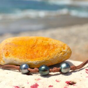 Full view of the leather bracelet with three 9.5 mm Tahitian pearls and knots displayed on a rock with a yellow stone by the sea