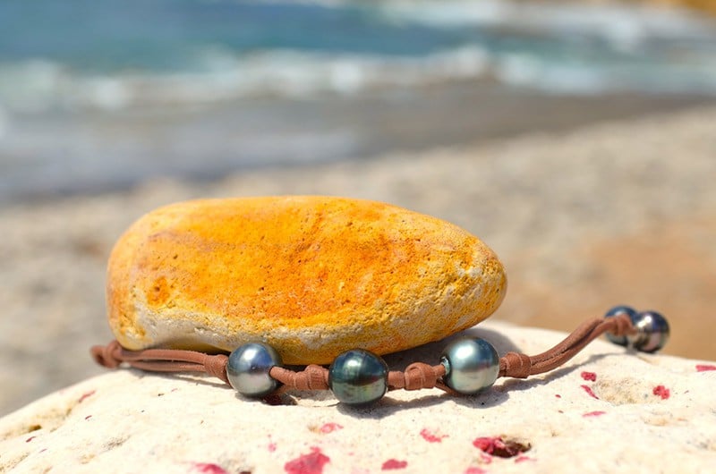 Full view of the leather bracelet with three 9.5 mm Tahitian pearls and knots displayed on a rock with a yellow stone by the sea