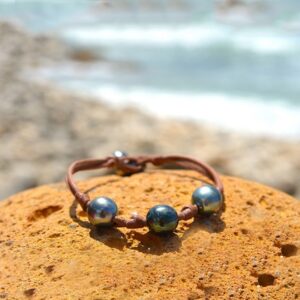 Brown leather bracelet with three central 9.5 mm Tahitian pearls, knots and two closure pearls displayed on a rock by the sea
