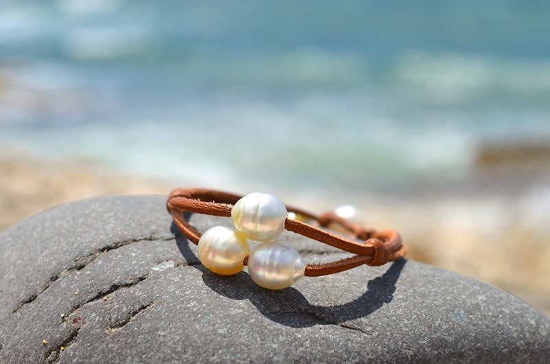 Bracelet en cuir marron à deux rangs avec trois perles blanches de culture d’Australie présenté sur une roche face à la mer