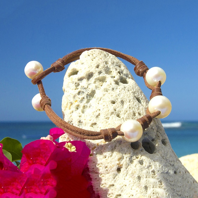 Bracelet en cuir avec trois perles blanches d’eau douce présenté autour d’une roche avec des fleurs roses face à la mer
