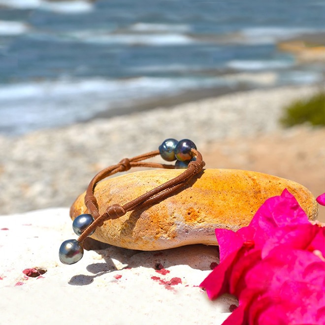 Bracelet en cuir marron à deux rangs avec trois perles baroques de Tahiti présenté sur une roche avec des fleurs roses
