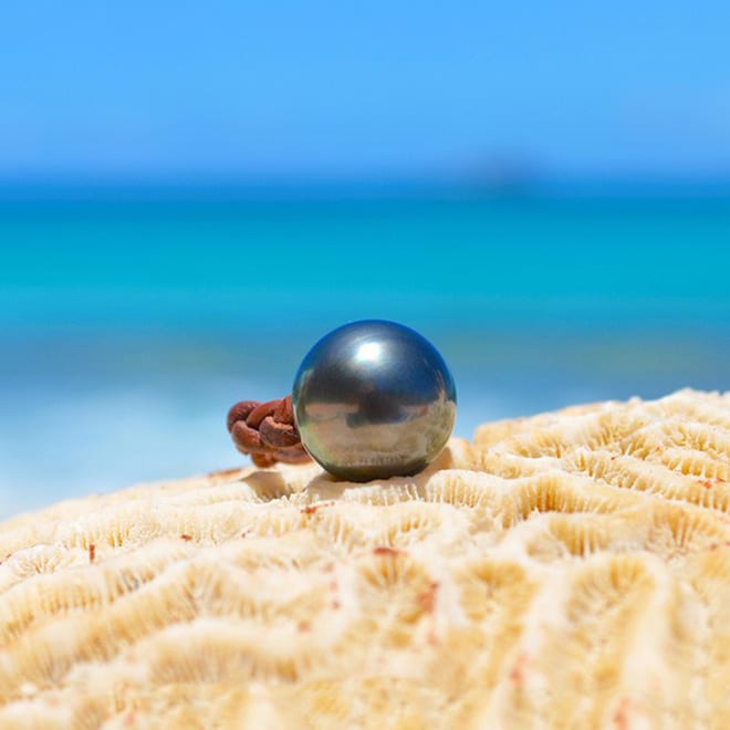 Bague en cuir marron tressé avec une perle de culture de Tahiti de 15 mm présentée sur du corail face à la mer