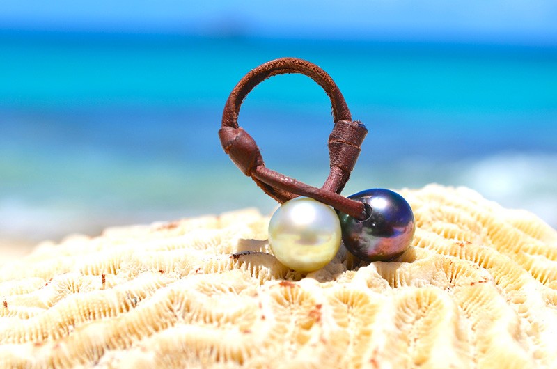 Bague Toi et Moi en cuir avec une perle de Tahiti et une perle blanche d’Australie de 11 mm présentée sur du corail face à la mer