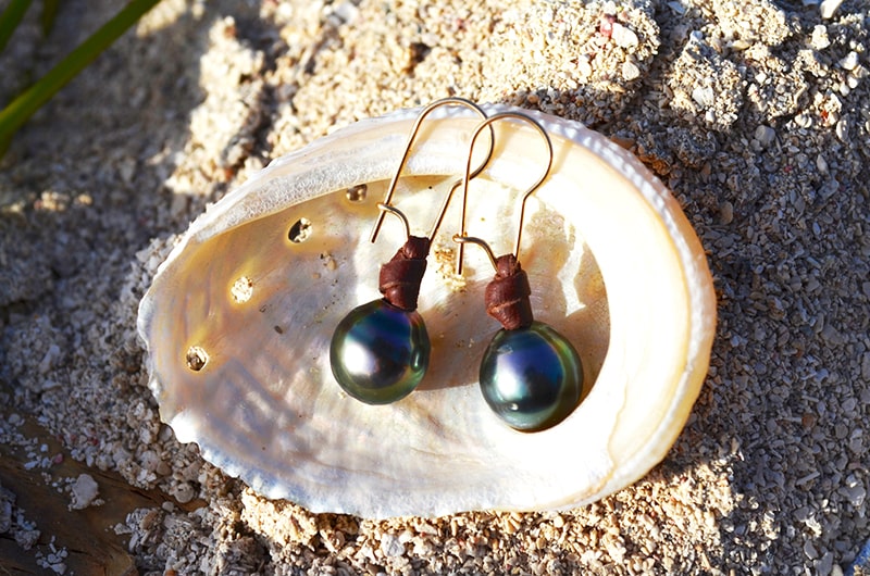 Leather earrings with semi-round Tahitian pearls displayed inside a seashell on a rock