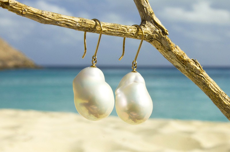 Gold earrings with white freshwater pearls suspended above a Saint Barthélemy beach