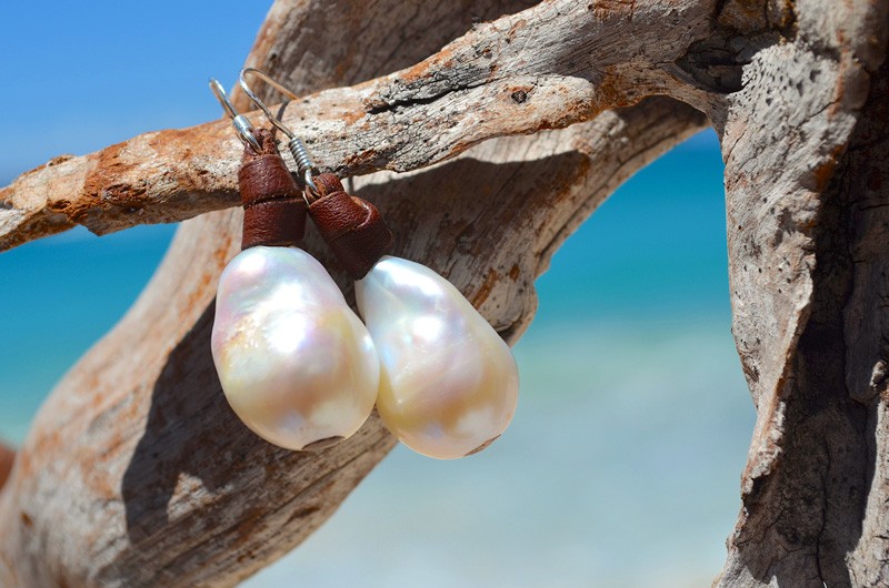 Leather earrings with white freshwater pearls displayed on driftwood by the sea