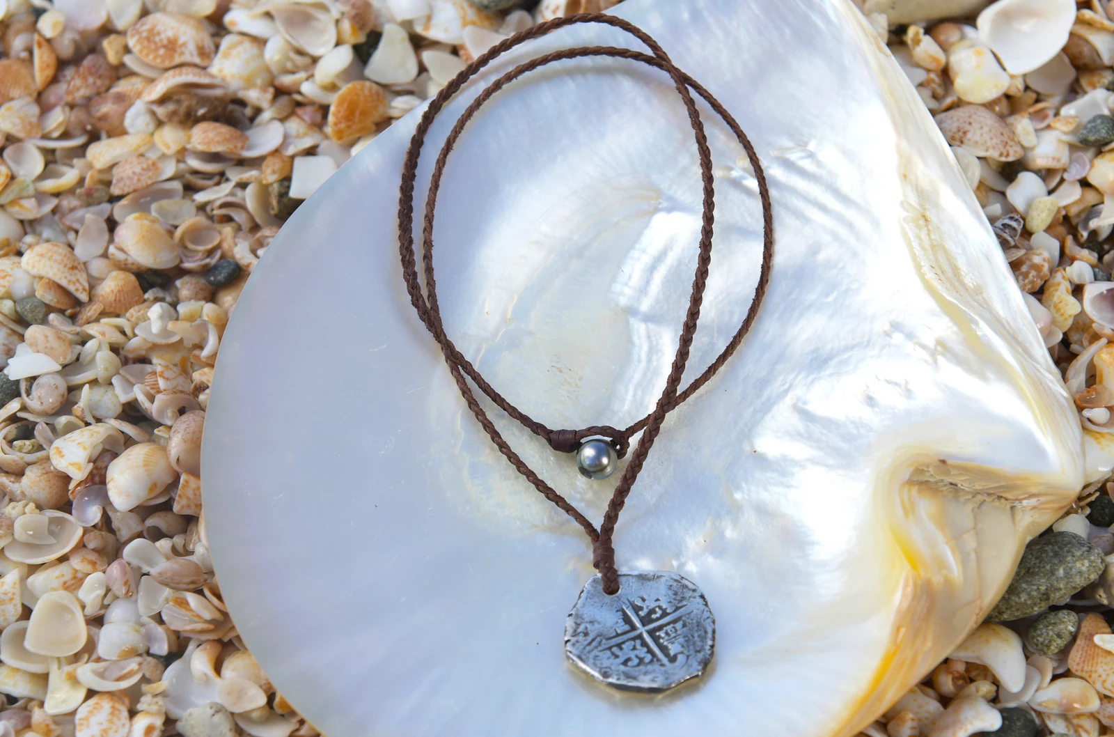 Braided leather pendant necklace with an antique silver eight reales coin and a Tahitian pearl displayed on mother-of-pearl