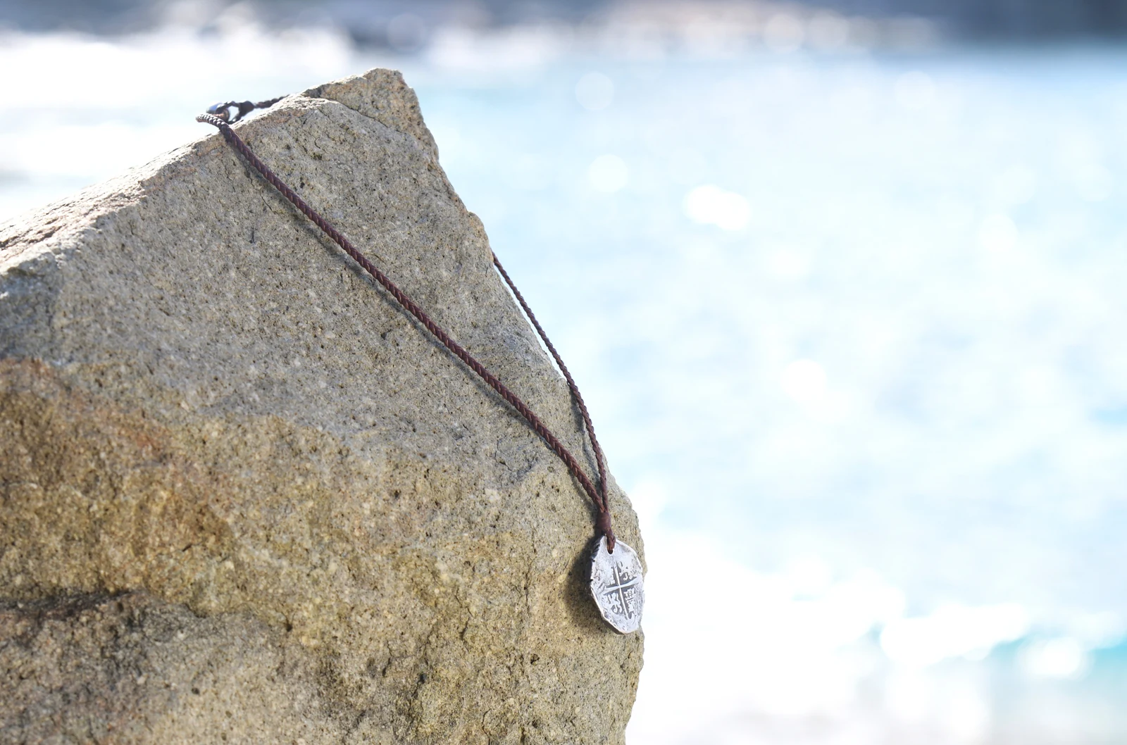 Braided leather pendant necklace with an antique silver eight reales coin displayed on a rock by the sea