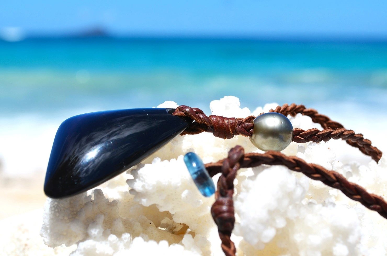 Vue latérale de l’obsidienne Œil Céleste, de la perle de Tahiti et du montage tressé du collier sur du corail blanc face à la mer