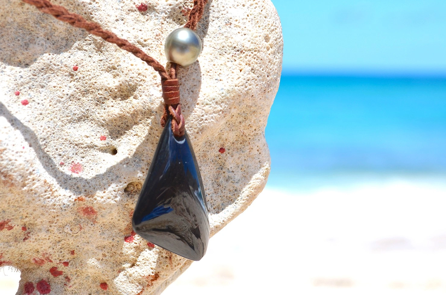Vue rapprochée du pendentif en obsidienne Œil Céleste et de la perle de culture de Tahiti du collier tressé face à la mer