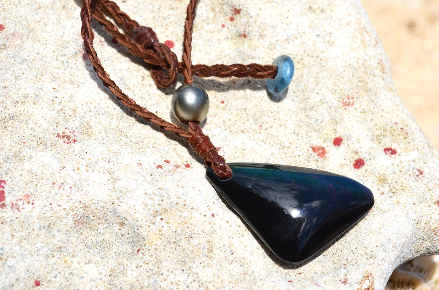 Détail de l’obsidienne Œil Céleste, de la perle de culture de Tahiti et du cuir tressé du collier présentés sur une roche claire