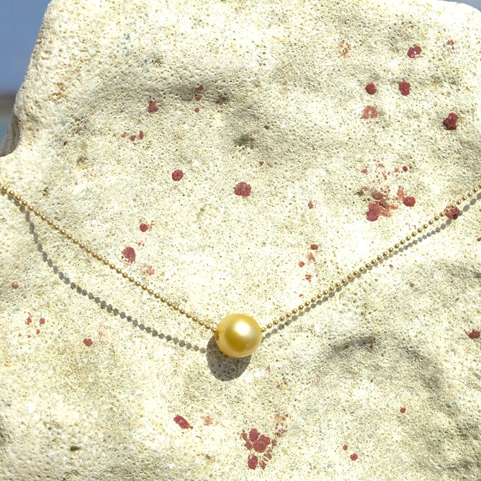 Full view of the 14-karat gold chain necklace with a 12 mm golden South Sea pearl displayed on coral facing the sea