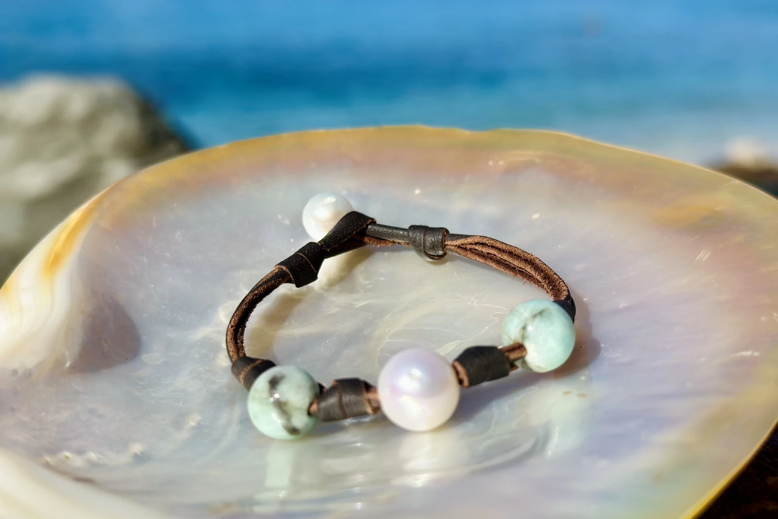 Bracelet en cuir marron avec billes d’émeraude et perle de culture blanche présenté sur une nacre face à la mer