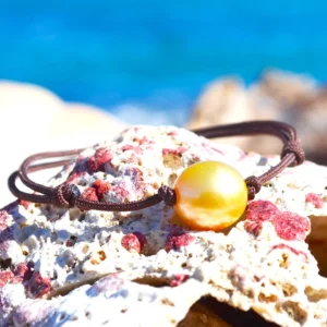 Bracelet ajustable sur cordon marron avec une perle de culture dorée d’Australie de 11,5 mm présenté sur une roche face à la mer