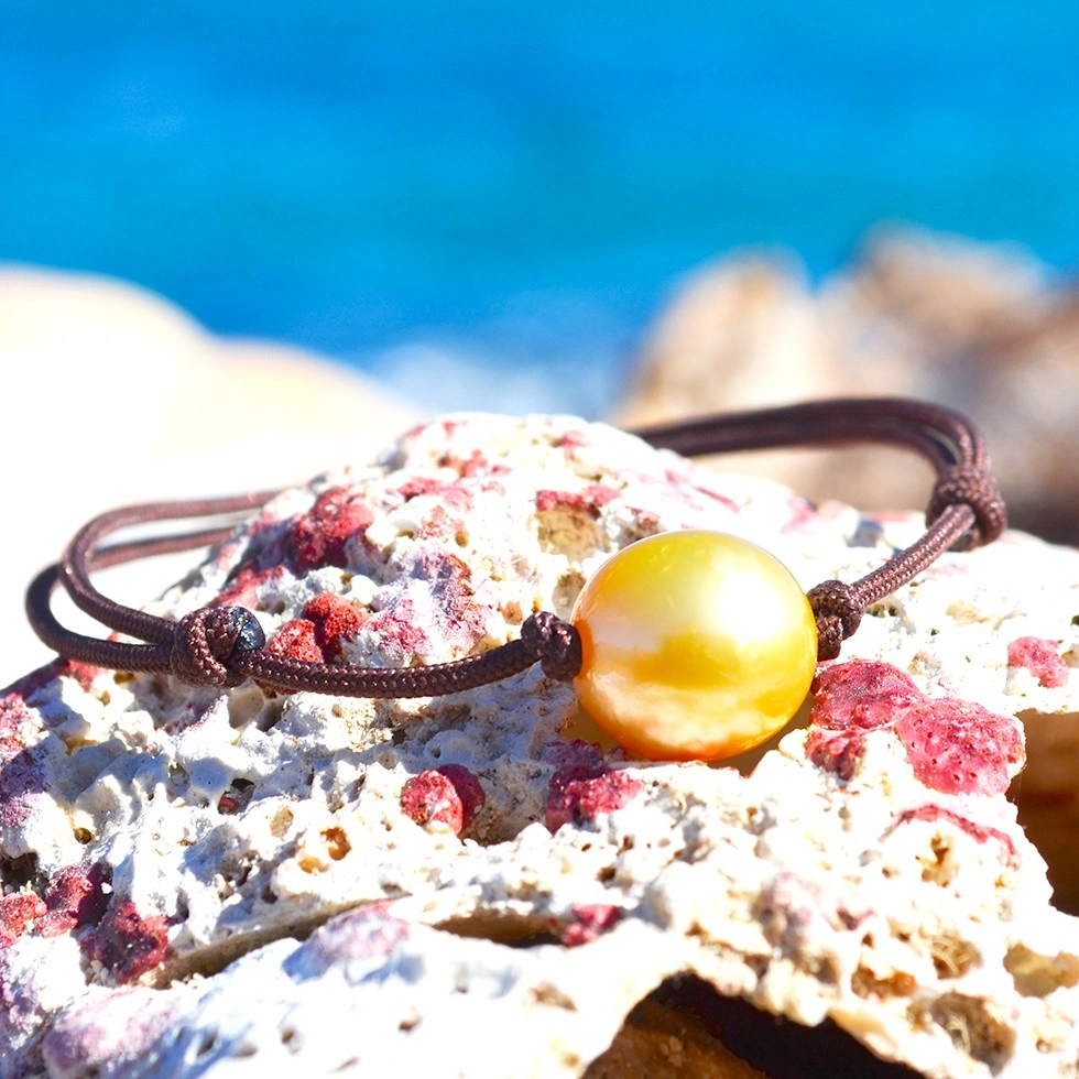 Bracelet ajustable sur cordon marron avec une perle de culture dorée d’Australie de 11,5 mm présenté sur une roche face à la mer