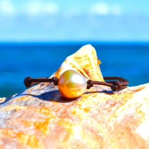Bracelet ajustable sur cordon marron avec une perle de culture dorée d’Australie de 12 mm présenté sur un coquillage face à la mer